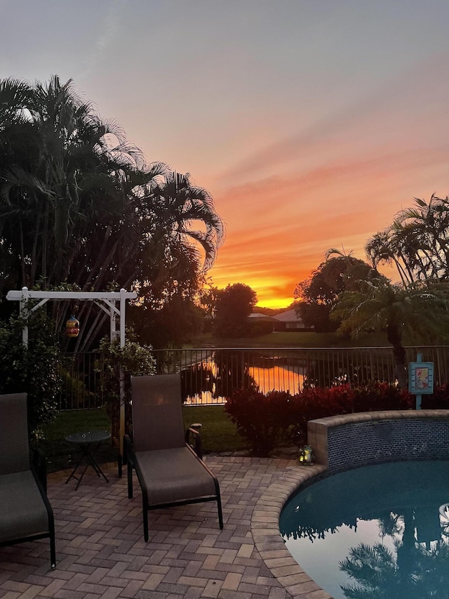 pool at dusk with a patio, a water view, fence, and a fenced in pool