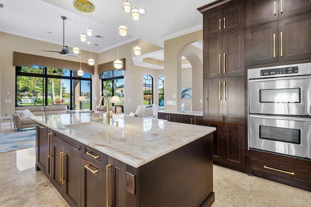 kitchen featuring sink, an island with sink, decorative light fixtures, light stone counters, and stainless steel double oven