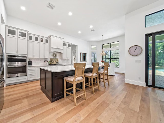 kitchen with light hardwood / wood-style floors, a center island with sink, backsplash, hanging light fixtures, and white cabinets