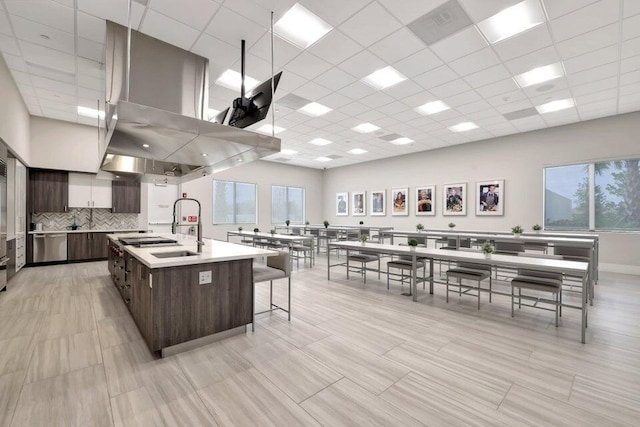 kitchen with island range hood, dark brown cabinetry, a center island with sink, backsplash, and a paneled ceiling