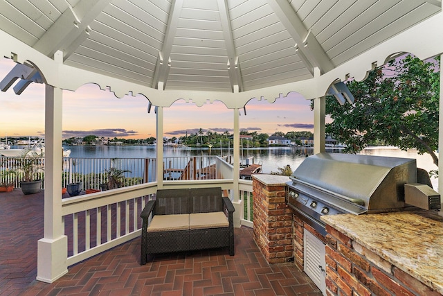 patio terrace at dusk with a gazebo, area for grilling, fence, and a water view