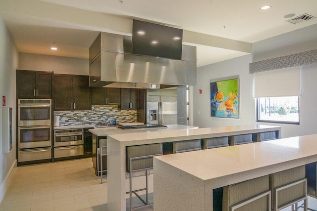 kitchen featuring a breakfast bar, backsplash, wall chimney exhaust hood, a large island, and stainless steel appliances