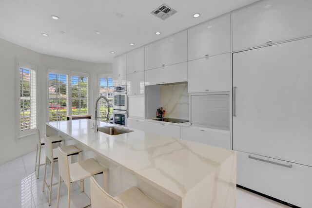 kitchen with black electric stovetop, a kitchen breakfast bar, stainless steel double oven, and an island with sink