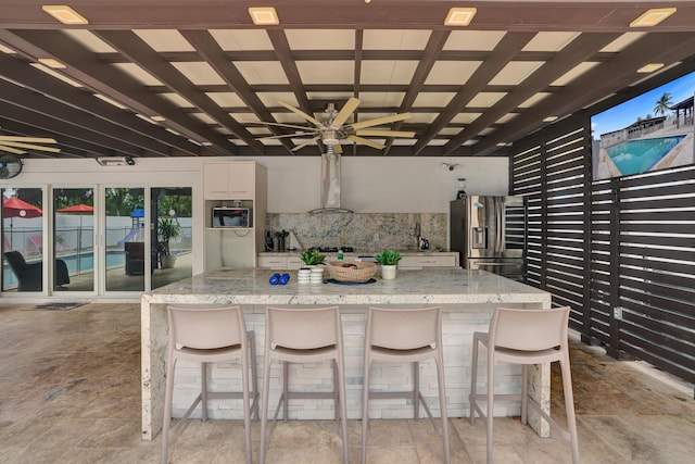 kitchen featuring white cabinetry, wall chimney exhaust hood, tasteful backsplash, stainless steel refrigerator with ice dispenser, and a kitchen bar