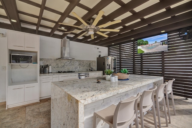 kitchen featuring light stone counters, wall chimney exhaust hood, stainless steel appliances, beamed ceiling, and white cabinetry