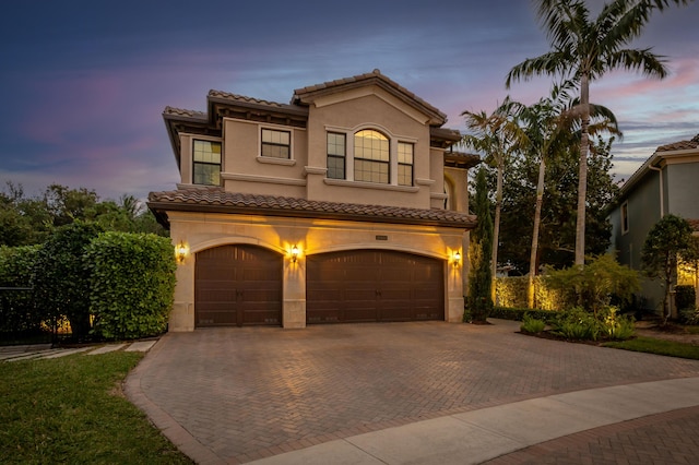 mediterranean / spanish-style house with an attached garage, a tiled roof, decorative driveway, and stucco siding