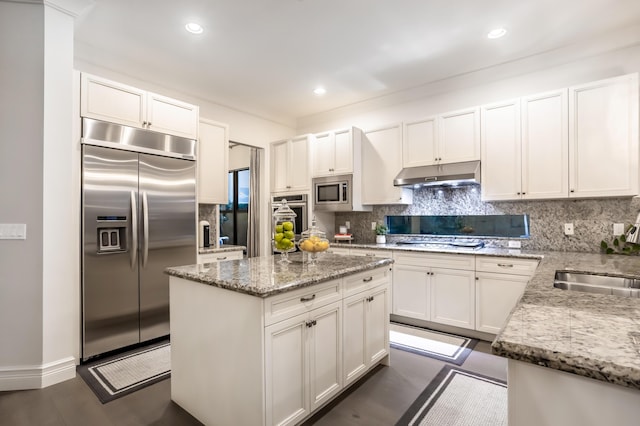 kitchen featuring light stone counters, built in appliances, under cabinet range hood, white cabinetry, and a sink