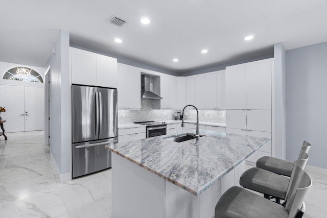 kitchen featuring sink, wall chimney exhaust hood, light stone countertops, a center island with sink, and appliances with stainless steel finishes