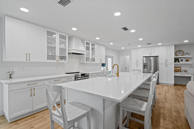 kitchen with stainless steel appliances, an island with sink, custom range hood, and white cabinetry