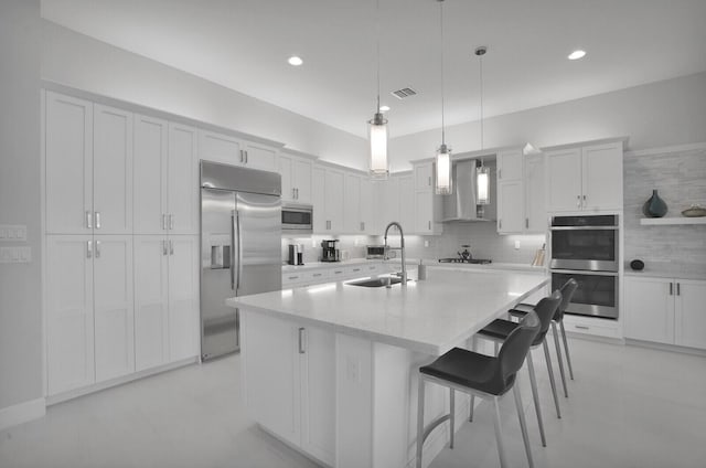 kitchen with sink, built in appliances, an island with sink, wall chimney range hood, and a breakfast bar