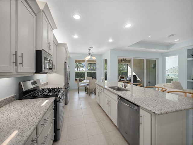 kitchen with a raised ceiling, sink, light stone counters, a center island with sink, and stainless steel appliances