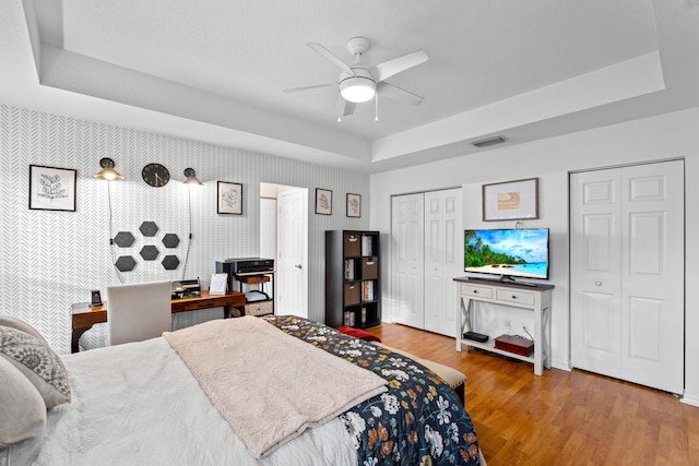 bedroom featuring ceiling fan, hardwood / wood-style floors, a tray ceiling, and multiple closets