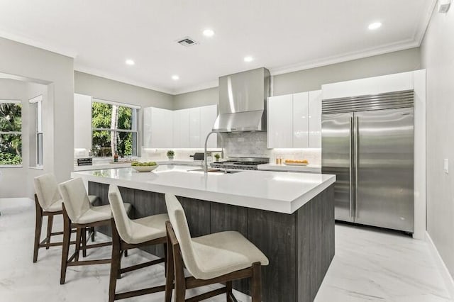 kitchen with stainless steel built in fridge, a center island with sink, white cabinets, and wall chimney exhaust hood