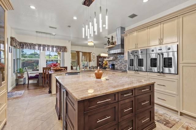 kitchen with light stone countertops, cream cabinets, wall chimney exhaust hood, hanging light fixtures, and dark brown cabinetry