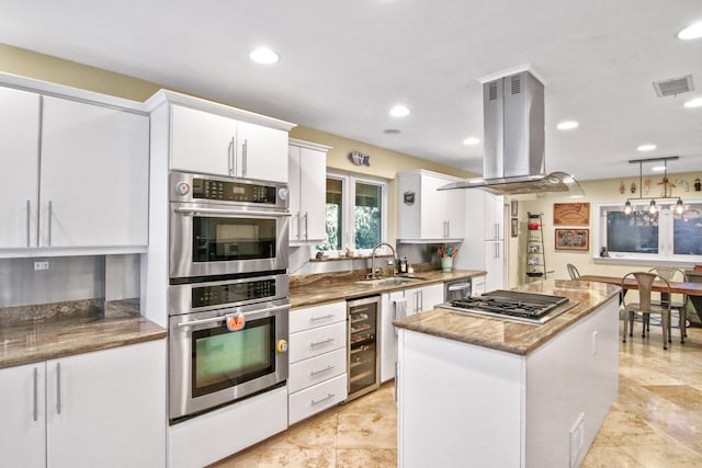 kitchen with island exhaust hood, appliances with stainless steel finishes, beverage cooler, sink, and white cabinetry