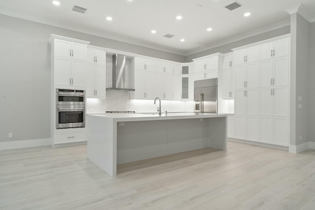 kitchen with sink, stainless steel appliances, wall chimney range hood, a center island with sink, and white cabinets