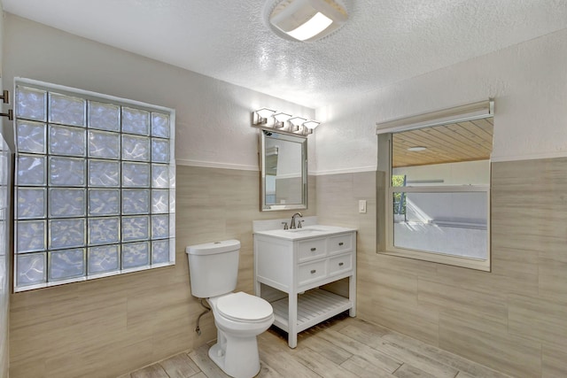 bathroom featuring a textured ceiling, a textured wall, toilet, vanity, and tile walls