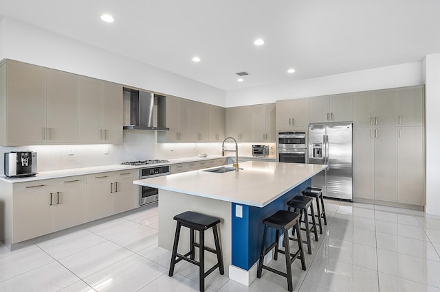 kitchen featuring wall chimney exhaust hood, stainless steel appliances, a kitchen island with sink, sink, and a breakfast bar area