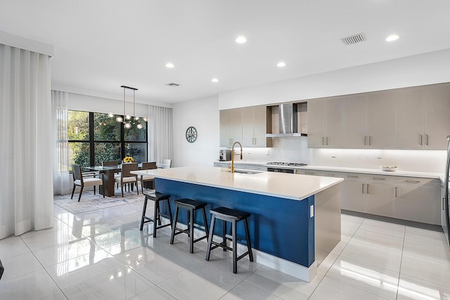 kitchen with wall chimney range hood, gray cabinets, an island with sink, a breakfast bar area, and stainless steel gas cooktop