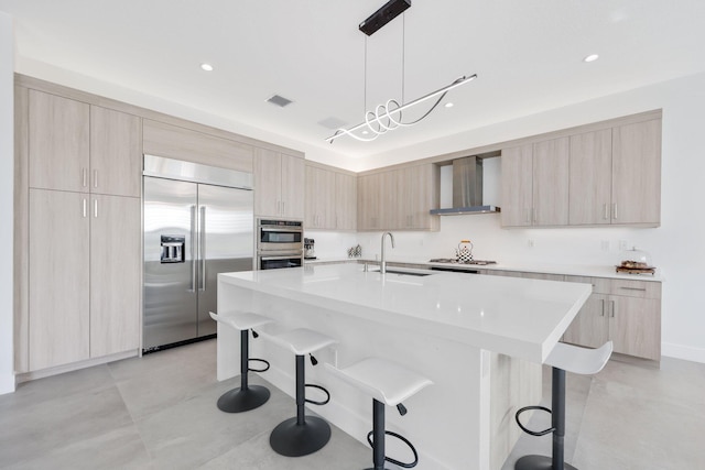 kitchen with appliances with stainless steel finishes, wall chimney exhaust hood, sink, decorative light fixtures, and an island with sink