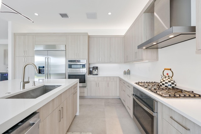 kitchen with light brown cabinets, sink, wall chimney range hood, pendant lighting, and appliances with stainless steel finishes