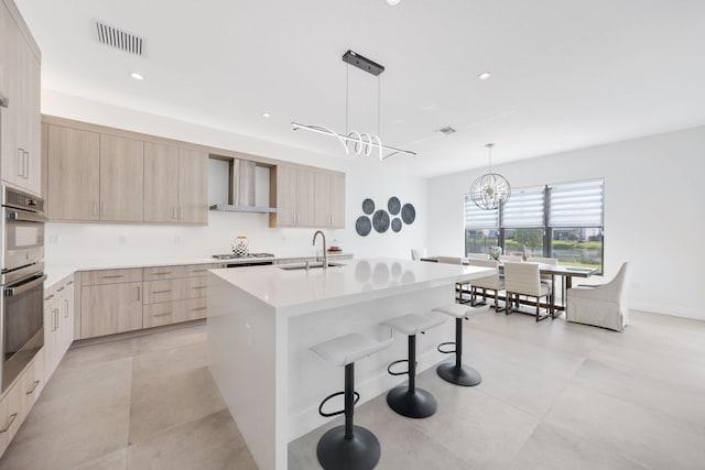 kitchen featuring sink, light brown cabinets, wall chimney range hood, pendant lighting, and a center island with sink