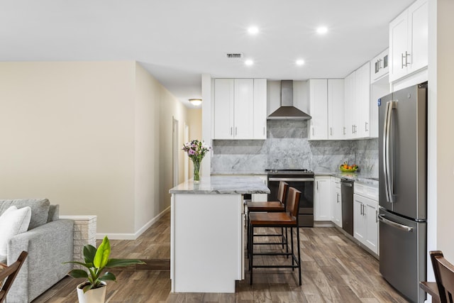 kitchen with appliances with stainless steel finishes, wall chimney exhaust hood, a kitchen island, white cabinetry, and a breakfast bar area