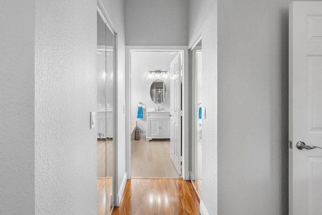 corridor featuring light wood finished floors, a textured wall, baseboards, and a sink