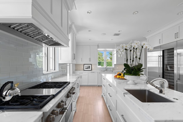 kitchen featuring decorative backsplash, premium range hood, light stone counters, sink, and white cabinetry