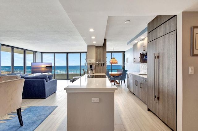 kitchen with paneled built in fridge, a wall of windows, decorative backsplash, hanging light fixtures, and a kitchen island with sink