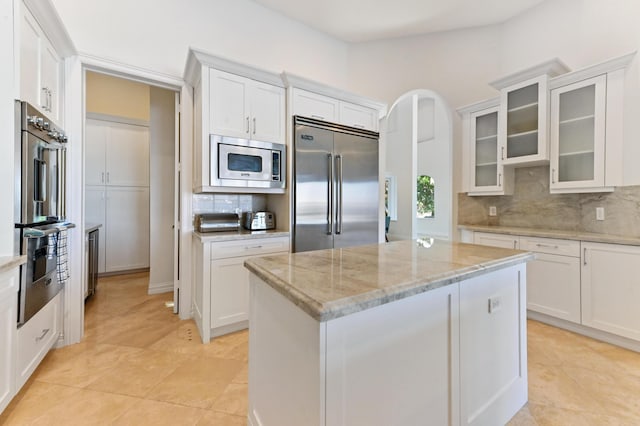 kitchen featuring light stone countertops, decorative backsplash, built in appliances, a center island, and white cabinetry