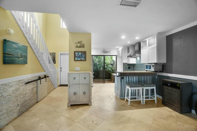 kitchen with wall chimney exhaust hood, a kitchen bar, white cabinetry, backsplash, and kitchen peninsula