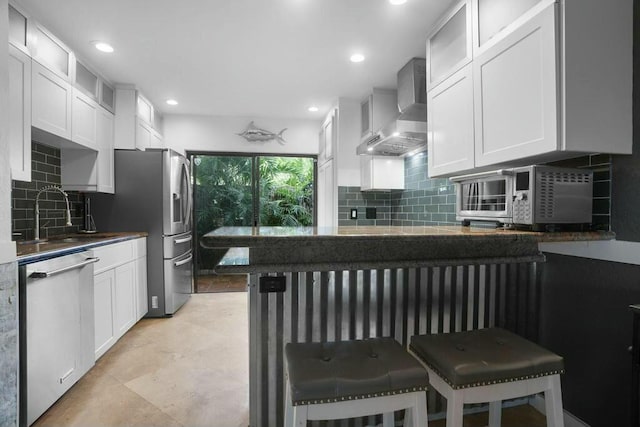 kitchen with a kitchen breakfast bar, wall chimney range hood, stainless steel appliances, and white cabinetry