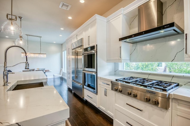 kitchen with appliances with stainless steel finishes, white cabinetry, hanging light fixtures, and wall chimney range hood