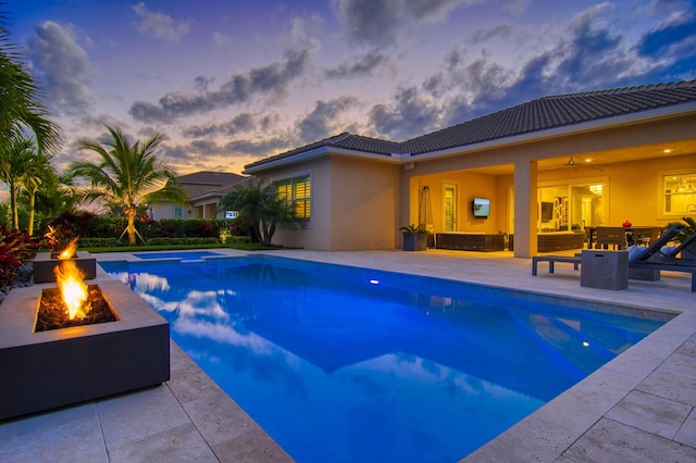 pool at dusk with ceiling fan, a patio, a fire pit, and an in ground hot tub