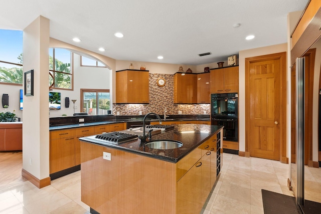 kitchen with sink, tasteful backsplash, black double oven, dark stone counters, and a kitchen island with sink