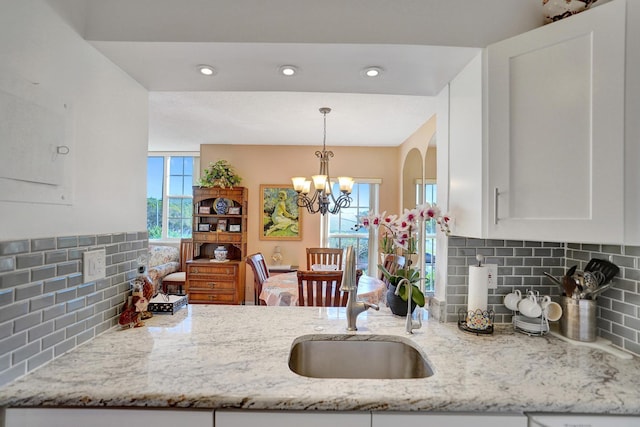 kitchen with tasteful backsplash, white cabinets, light stone countertops, an inviting chandelier, and a sink