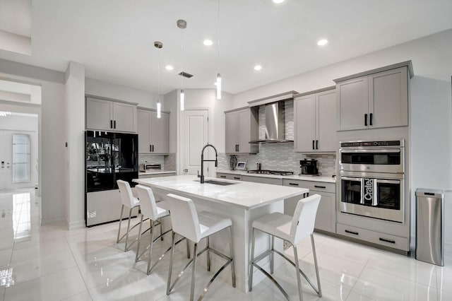 kitchen with stainless steel appliances, an island with sink, hanging light fixtures, sink, and wall chimney range hood