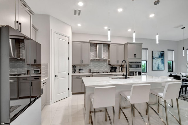 kitchen featuring sink, wall chimney exhaust hood, an island with sink, hanging light fixtures, and gray cabinetry