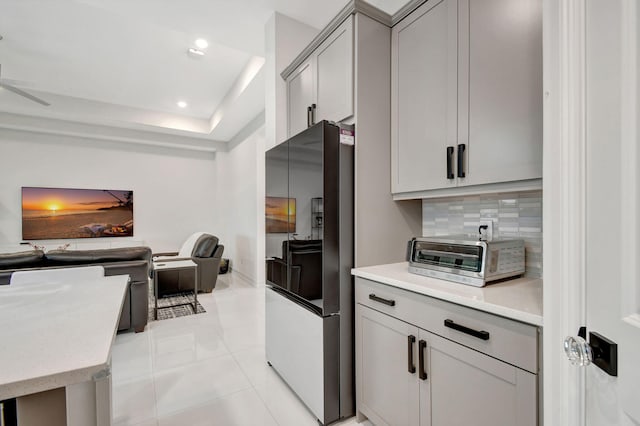 kitchen with black refrigerator, gray cabinets, light tile patterned floors, decorative backsplash, and a tray ceiling