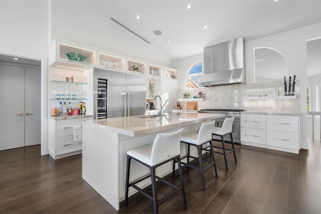 kitchen with high end appliances, wall chimney range hood, white cabinets, a breakfast bar, and a center island with sink