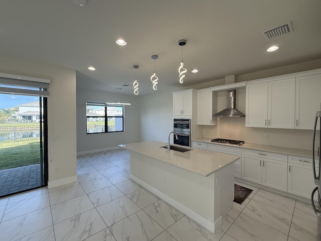 kitchen featuring light stone countertops, wall chimney exhaust hood, hanging light fixtures, an island with sink, and white cabinets