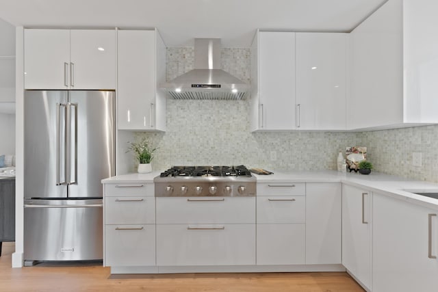 kitchen with wall chimney range hood, white cabinetry, stainless steel appliances, light hardwood / wood-style floors, and decorative backsplash