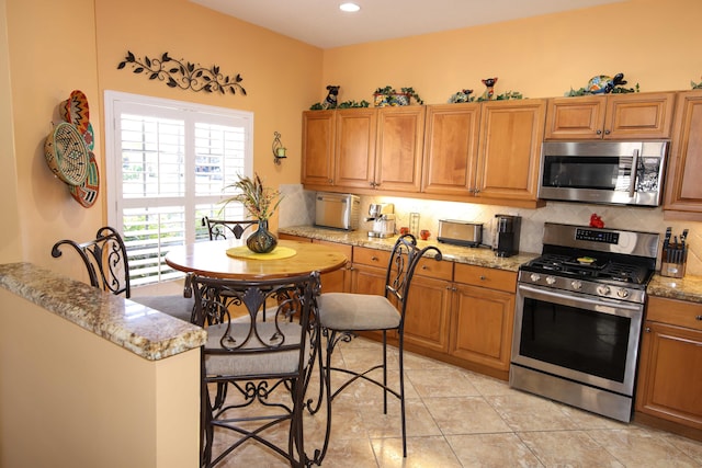 kitchen featuring a kitchen bar, light tile patterned floors, backsplash, light stone countertops, and appliances with stainless steel finishes