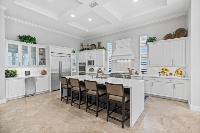 kitchen with premium range hood, a kitchen island with sink, crown molding, built in appliances, and white cabinets