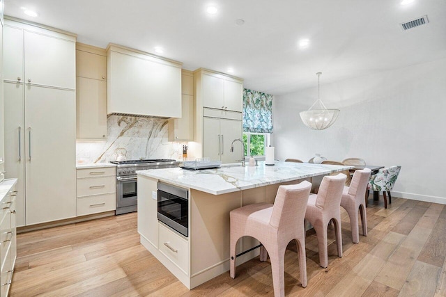 kitchen with light hardwood / wood-style flooring, built in appliances, cream cabinets, an island with sink, and decorative backsplash