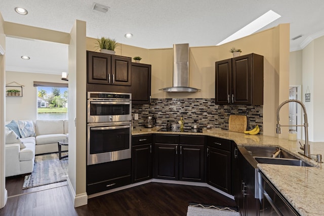 kitchen featuring light stone counters, black electric cooktop, stainless steel double oven, wall chimney range hood, and backsplash