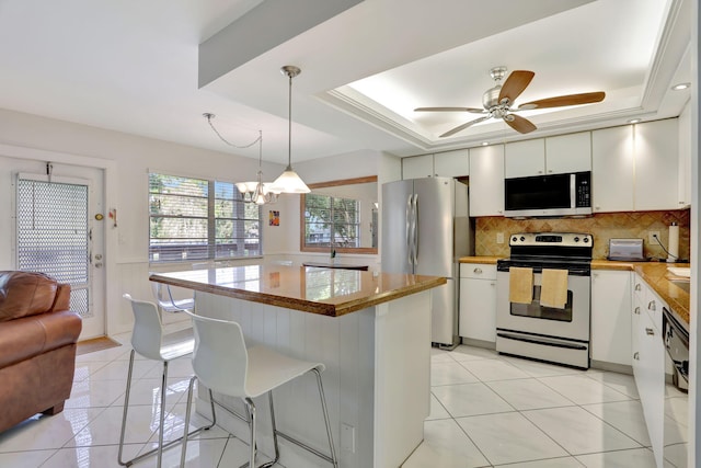 kitchen featuring a raised ceiling, stainless steel appliances, white cabinets, decorative light fixtures, and backsplash