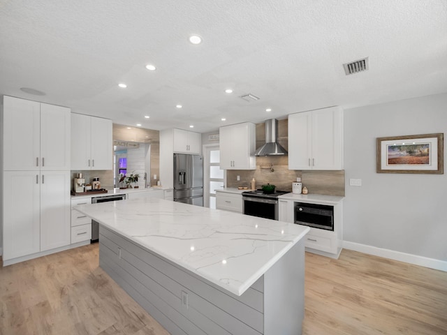 kitchen featuring light stone countertops, wall chimney exhaust hood, stainless steel appliances, a center island, and white cabinetry