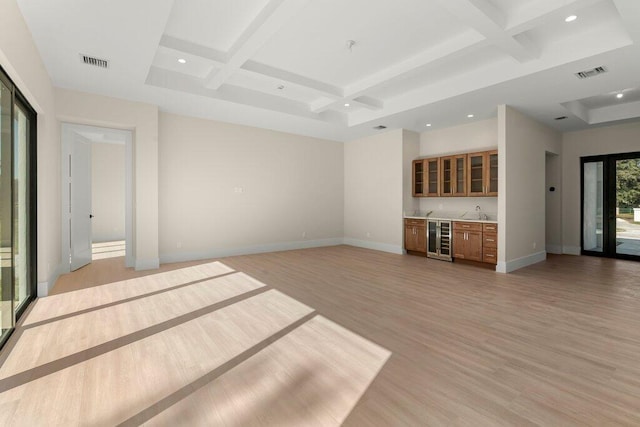 unfurnished living room featuring coffered ceiling, light wood-type flooring, indoor wet bar, wine cooler, and beamed ceiling